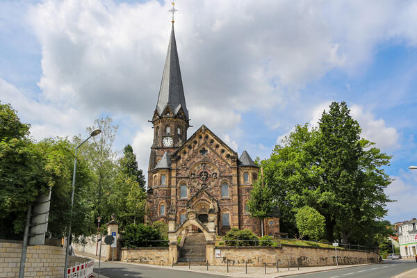 Vorderansicht der D&ouml;hlener Lutherkirche. Diese ist mit dunklen Steinen erbaut und weist an der linken Seite einen Spitzen Turm mit einer Uhr auf. Links und rechts der Kirche stehen B&auml;ume und im Vordergrund ist ein Teil der Lutherstra&szlig;e zu erkennen. &Uuml;ber der Kirche ist eine gro&szlig;e wei&szlig;e Wolke.