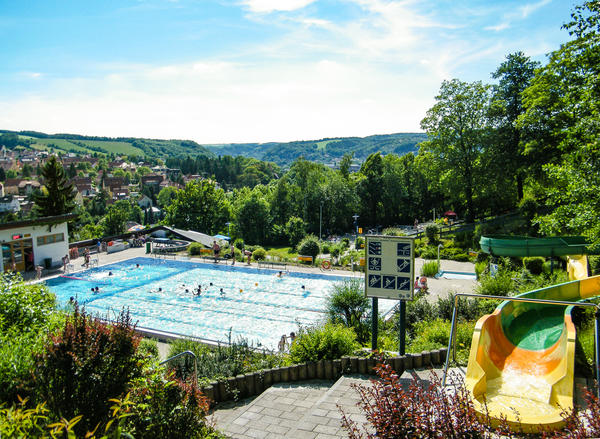 Blick von einer Anh�he auf ein Schwimmbecken, in welchem sich Personen befinden. Neben dem Schwimmbecken ein wei�es Haus. Auf die Anh�he f�hrt eine Treppe. Neben der Treppe befinden sich Bepflanzungen. Auf einer der bepflanzten Fl�chen steht ein Hinweisschild mit Piktogrammen. Daneben befindet sich der Einstieg zu einer bunten Rutsche, welche teilweise auch weiter im Hintergrund noch zusehen ist. Hinter dem Freibad sind B�ume zusehen. Auf der rechten hinteren Bildseite sind zur Stadt geh�rige Siedlungen zu erkennen. Im Hintergrund erheben sich bewaldete Anh�hen vor hellem Himmel.
