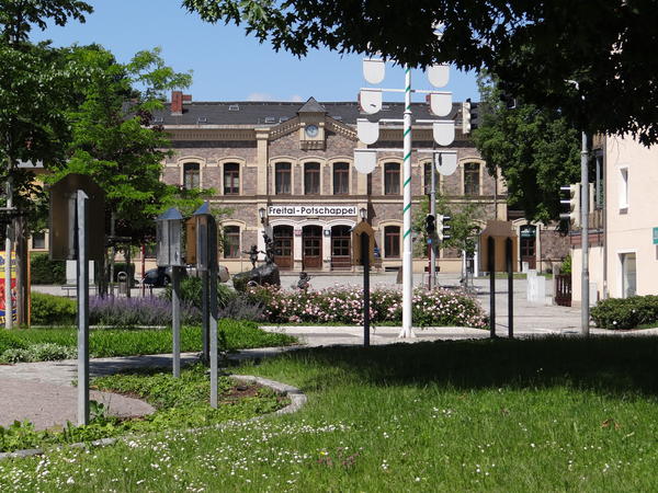 Das Bild zeigt im Hintergrund das Bahnhofsgeb�ude von Potschappel �ber dessen Eingang die Worte Freital-Potschappel stehen. Davor befinden sich ein Blumenbeet, Rasen und B�ume. Au�erdem ist ein Teil des Marktplatzes und ein dort befindliches Wasserspiel mit kleinen Figuren abgebildet.