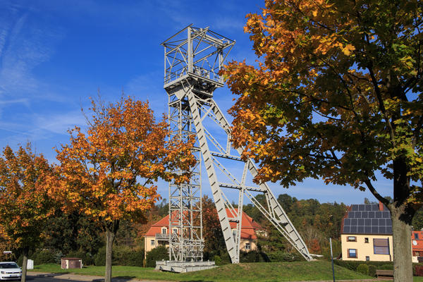 Das Bild zeigt ein F�rderger�st eines ehemaligen Bergbaubetriebes, welches auf einer Wiese steht. Vor dem Ger�st befinden sich herbstlich gef�rbte B�ume. Neben einem Baum parkt ein wei�es Auto, wobei das Bild nur dessen Frontbereich zeigt. Im Hintergrund  befinden sich drei gelbe H�user mit roten D�chern, wobei das mittelste sehr viele Solarpanels auf dem Dach aufweist. Hinter den H�usern ist Wald zu erkennen.