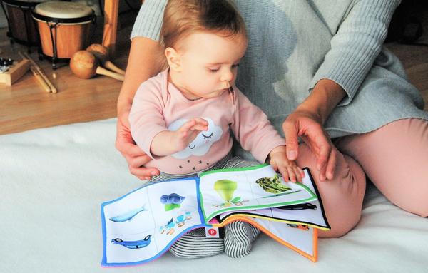 Ein kleines Kind in einem rosafarbenen Pullover sitzt auf einer wei�en Decke. In der Hand hat es ein buntes Bilderbuch. Neben dem Kind ist der Unterk�rper einer Person in einer rosafarbenen Hose und mit einem grau-gr�nen Pullover zusehen, welche das Kind festh�lt. Im Hintergrund liegen verschiedene Musikinstrumente auf Parkettboden.