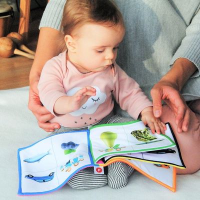 Ein kleines Kind in einem rosafarbenen Pullover sitzt auf einer wei&szlig;en Decke. In der Hand hat es ein buntes Bilderbuch. Neben dem Kind ist der Unterk&ouml;rper einer Person in einer rosafarbenen Hose und mit einem grau-gr&uuml;nen Pullover zusehen, welche das Kind festh&auml;lt. Im Hintergrund liegen verschiedene Musikinstrumente auf Parkettboden.