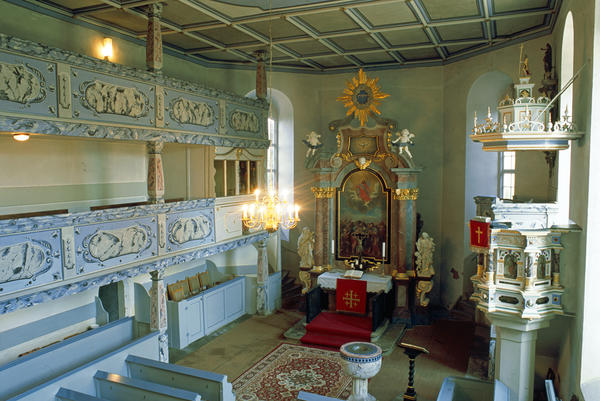 Blick in das Innere einer Kirche. Zusehen sind die wei&szlig;-blau gehaltenen Emporen sowie ein Teil der B&auml;nke. Dahinter der mit goldenen Elementen versehene Altar mit dem davor befindlichen roten Teppich sowie das davor befindliche Taufbecken. Rechts die helle Kanzel.