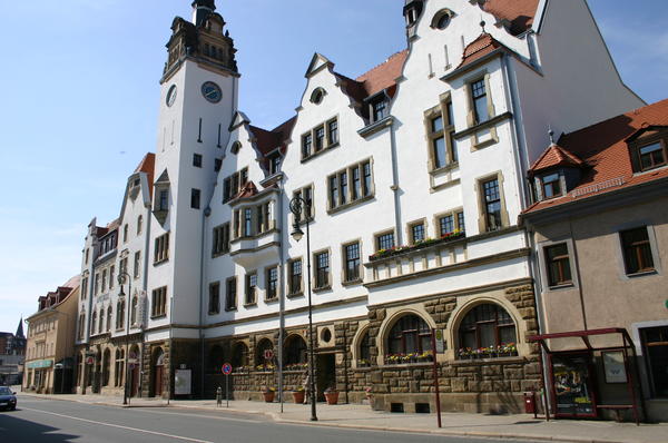 Zu sehen ist die Vorderseite des Rathauses in Potschappel und ein Teil der Dresdner Stra�e. Das Rathaus ist ein wei�es, mehrst�ckiges Geb�ude mit vielen kleinen Fenstern und einem spitzen Turm in den eine Uhr eingelassen ist. Am rechten unteren Bildrand ist zudem die Bushaltestelle, welche sich vor dem Rathaus befindet, abgebildet.