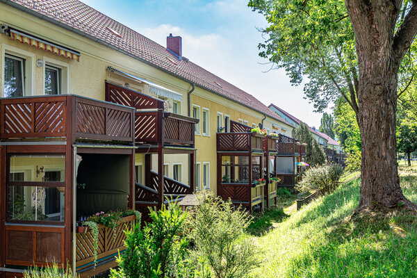 Das Bild zeigt ein gelb-oranges Mehrfamilienhaus mit braunen Balkonen davor. Auf der rechten Seite befindet sich eine B&ouml;schung mit einem Baum.