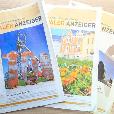 Drei Ausgaben der Zeitung Freitaler Anzeiger liegen einander &uuml;berlappend auf einer Oberfl&auml;che. Alle haben relativ mittig ein gro&szlig;es Bild. Auf der obersten Zeitung ist sowohl unten, als auch am Rand Text zusehen. zudem ist in der linken unteren Ecke das Logo Freitals zusehen.