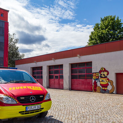 Das Foto zeigt einen roten Transporter, auf dessen Motorhaube "Feuerwehr" steht. Dieser befindet sich auf einem gepflasterten Hof. Hinter dem Auto ist ein roter Turm zusehen. Direkt daneben befindet sich ein flaches, helles Geb�ude mit roten Toren. An eine Stelle des Geb�udes sind mit Graffiti das Logo der Stadt Freital und direkt daneben ein Feuerwehrmann mit einem Wasserschlauch in der Hand aufgespr�ht. Im oberen Bildteil sind die Krone eines Baumes und teilweise bew�lkter Himmel zusehen.