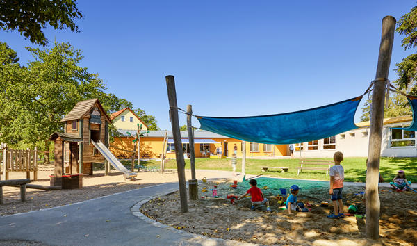Fotografiert ist ein von einem Weg durchzogener Au�enbereich. Auf der rechten Seite, neben dem Weg, befindet sich ein mit einem blauen Sonnensegel �berdachter Sandkasten, in welchem Kinder spielen. Auf der linken Seite ist ein Spielhaus mit einer Rutsche. Im Hintergrund steigt eine Wiese leicht an, dahinter stehen zwei flache, helle Geb�ude. An den Bildseiten und im Hintergrund befinden sich B�ume. Im oberen Bildteil zeigt sich ein wolkenloser Himmel.