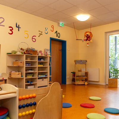 Das Bild zeigt einen Raum, welcher mit Holzm&ouml;beln ausgestattet ist. In einem Regal an der Wand befinden sich verschiedene Dinge. &Uuml;ber dem Regal sind bunte Zahlen an einer gelben Wand angebracht. Rechts daneben ist eine Holzt&uuml;r. &Uuml;ber dieser h&auml;ngt eine kleine Schaukel, auf welcher eine Stoffpuppe sitzt. Auf dem Boden liegen bunte Kissen. Im Hintergrund befindet sine offene Glast&uuml;r, welche ins freie f&uuml;hrt.