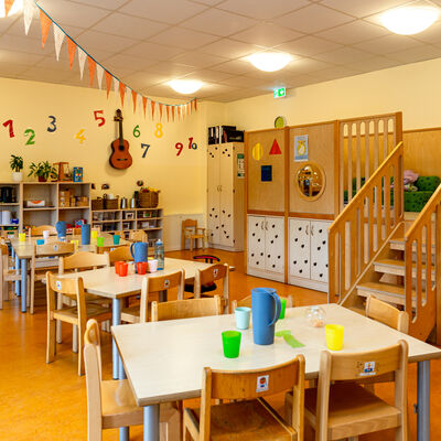 Blick in einen Raum mit kleinen Holztischen und St&uuml;hlen. Auf den Tischen stehen bunte Becher. An der Decke sind Wimpel befestigt. An der Wand im Hintergrund befindet sich ein Regal mit einigen Dingen darin. Dar&uuml;ber kleben bunte Buchstaben an der Wand, zwischen denen eine Gitarre h&auml;ngt. Auf der rechten Bildseite ist ein kleines mit einer Treppe versehenes Spielepodest.
