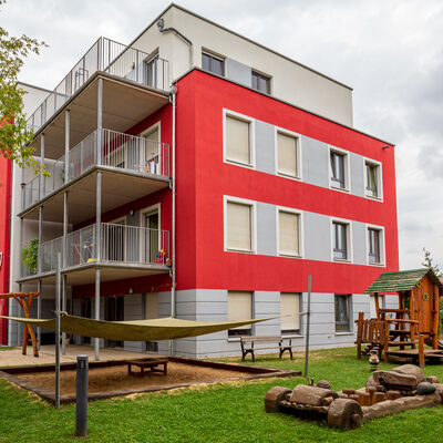 Blick auf ein mehrst&ouml;ckiges, rotes Geb&auml;ude mit grauen und wei&szlig;en Elementen. Vor dem Geb&auml;ude befindet sich ein aus Holz gebauter Spielbereich auf einer Wiese. Daneben ein Sandkasten, &uuml;ber welchem ein helles Tuch gespannt ist. Neben dem Sandkasten verl&auml;uft ein Weg. Hinter dem Sandkasten ist eine auf einem Sandplatz errichtete Netzschaukel zu erkennen. Im oberen Bildteil ist bedeckter Himmel.