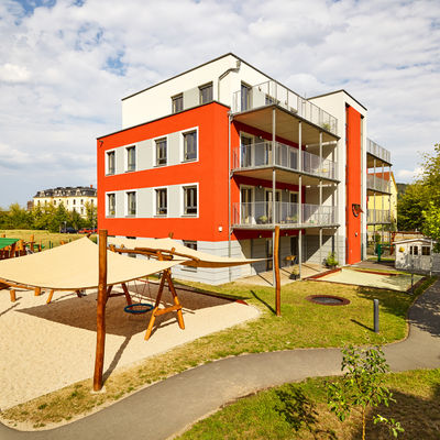Blick auf ein mehrst&ouml;ckiges, rotes Geb&auml;ude mit grauen und wei&szlig;en Elementen. Vor dem Geb&auml;ude befindet sich ein aus Holz gebauter Spielbereich auf einem Sandplatz, &uuml;ber welchem ein helles Tuch gespannt ist. Neben dem Sandplatz verl&auml;uft ein Weg, neben welchem sich eine mit B&auml;umen versehene Rasenfl&auml;che befindet. Im Hintergrund weitere Geb&auml;ude und dar&uuml;ber wolkiger Himmel.