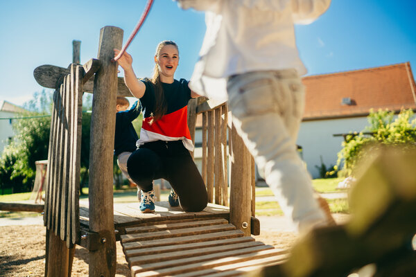 Zusehen ist der obere Teil eines h�lzernen Kletterturms, auf welchem eine dunkel angezogene Frau hockt und zu einem Kind schaut, dessen untere H�lfte ebenfalls auf dem Bild zusehen ist und welches, �ber eine h�lzerne Br�cke, auf die Frau zukommt. Im Hintergrund sind B�ume, Fassaden und wolkenloser Himmel.