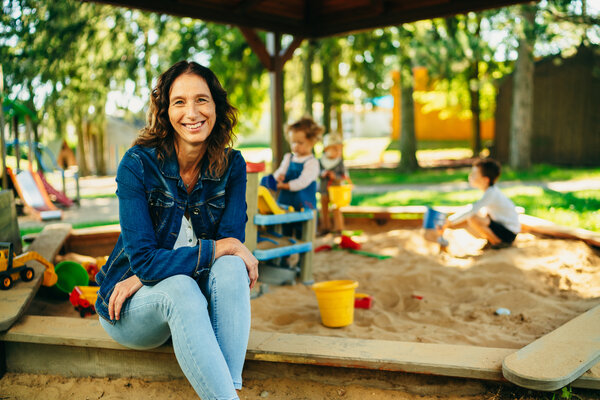 Das Bild zeigt eine blau angezogene Frau, welche auf dem Rand eines �berdachten Sandkastens sitzt und in die Kamera l�chelt. Hinter ihr spielen Kinder mit buntem Spielzeug im Sand. Im Hintergrund befinden sich weitere Spielm�glichkeiten sowie B�ume und Wiese.