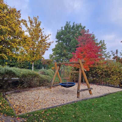 Auf einer Sandfl&auml;che steht ein Holzger&uuml;st mit einer Schaukel. Im Hintergrund herbstlich bunt gef&auml;rbte B&auml;ume.