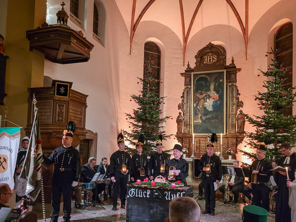 Zu sehen ist der Altarraum einer Kirche. Vor dem Altar, hinter dem sich ein kunstvoll golden umrahmtes Bild befindet, stehen mehrere Personen in dunklen, bergmanntypischen Sachen. Sie haben Laternen vor sich.  Links ist noch die Empore und im Hintergrund zwei Weihnachtsb&auml;ume und Kirchenfenster zu sehen.