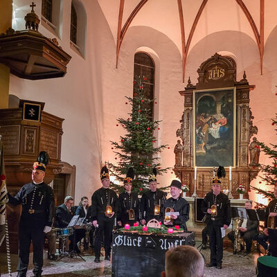 Zu sehen ist der Altarraum einer Kirche. Vor dem Altar, hinter dem sich ein kunstvoll golden umrahmtes Bild befindet, stehen mehrere Personen in dunklen, bergmanntypischen Sachen. Sie haben Laternen vor sich.  Links ist noch die Empore und im Hintergrund zwei Weihnachtsb&auml;ume und Kirchenfenster zu sehen. 