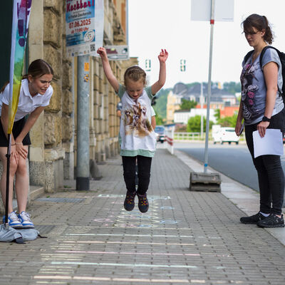 Kinder spielen auf dem Gehweg vor einem Gesch&auml;ft. Daneben fahren Autos auf der Stra&szlig;e.