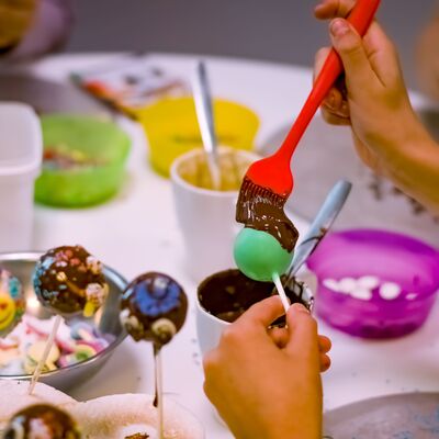 Kinderh&auml;nde dekorieren an einem Tisch Cakepops mit Schokolade und verschiedenen bunten Streuseln, die in Schalen auf dem Tisch verteilt sind