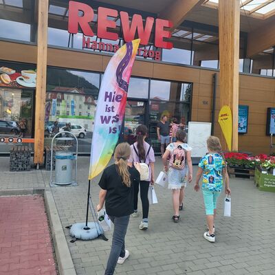 Vier Kinder laufen mit Papiert&uuml;ten in den H&auml;nden in einen gro&szlig;en Rewe-Supermarkt. Vor diesem stehen zwei Erwachsene, Pflanzen, ein Papierkorb und eine Beachflag mit der Aufschrift "Hier ist was los".