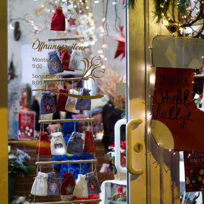 Die T&uuml;r und das Schaufenster eines Blumenladens sind mit einem gro&szlig;en Stiefel, Lichterketten, Tannengr&uuml;n und einem S&auml;ckchen-Adventskalender weihnachtlich dekoriert. Alles funkelt im Lichterglanz.