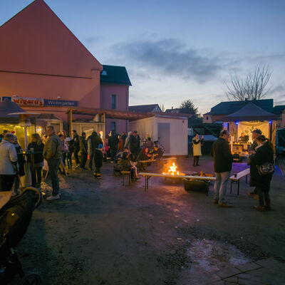 Es ist Abend. Auf einem kleinen Platz versammeln sich mehrere Menschen an St&auml;nden, Biertischen und einer Feuerschale, in der ein Feuer prasselt. Im Hintergrund sind gro&szlig;e H&auml;user zu sehen.