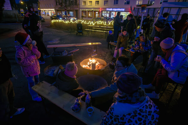Um eine Feuerschale stehen und sitzen Menschen auf Bierb&auml;nken, die Kn&uuml;ppelkuchen machen und essen. Im Hintergrund befinden sich eine Lichterkette an einer Hecke und die beleuchtete Stadt-Apotheke.