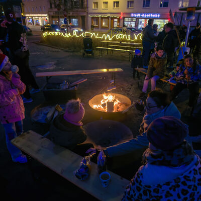 Um eine Feuerschale stehen und sitzen Menschen auf Bierb&auml;nken, die Kn&uuml;ppelkuchen machen und essen. Im Hintergrund befinden sich eine Lichterkette an einer Hecke und die beleuchtete Stadt-Apotheke.