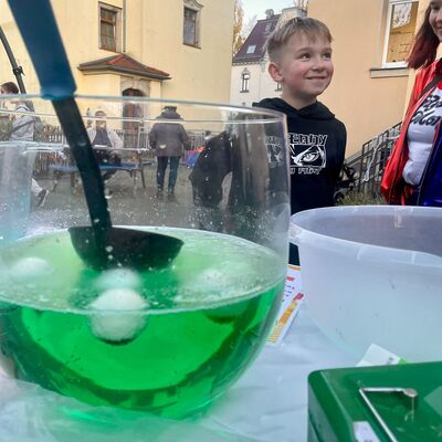 Auf einem Tisch steht eine gr&uuml;ne Gruselbowle. Darin schwimmen falsche Aug&auml;pfel. Im Hintergrund befinden sich ein Innenhof zwischen H&auml;usern mit einem Jungen und einem M&auml;dchen, die sich freuen.