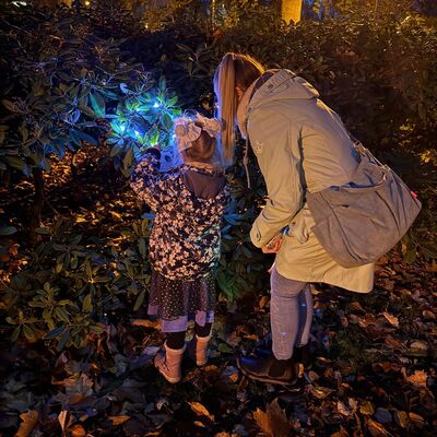 Es ist Nacht. Eine Frau und ein gruselig verkleidetes M&auml;dchen, die von hinten zu sehen sind, sehen sich eine Aufgabe des Gruselparcours an einer Hecke an. Auf dem Boden liegt altes Laub. Im Hintergrund beleuchtet eine Stra&szlig;enlaterne einige B&auml;ume.