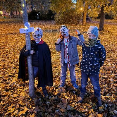 Es ist Nacht In einem gro&szlig;en Park stehen drei gruselig verkleidete und geschminkte Kinder an einem kleinen Baum. Daran h&auml;ngen eine Lichterkette und ein Zettel mit Aufgaben f&uuml;r die Kinder. Auf dem Boden liegt viel altes Laub. Im Hintergrund stehen gro&szlig;e B&auml;ume.