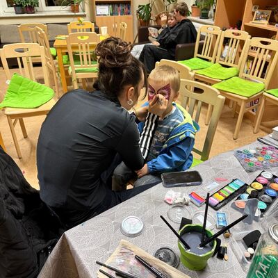 In einem Raum sitzt ein Kind auf einem Stuhl. Eine Frau schminkt das Kind. Davor steht ein Tisch mit vielen Utensilien f&uuml;r das Kinderschminken. Dahinter sitzen ein Kind und ein Erwachsener auf St&uuml;hlen mit gr&uuml;nen Kissen. Weitere St&uuml;hle sind im Raum verteilt. An der Wand stehen B&uuml;cherregale.