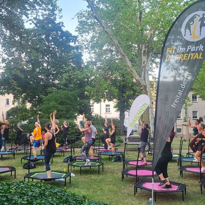 Es ist Sommer. Auf einer von B&uuml;schen und B&auml;umen umgebenen Wiese trainieren viele Menschen gemeinsam auf bunten Trampolinen. Dazwischen stehen Beachflags von Fit im Park und der Barmer-Krankenkasse. Im Hintergrund stehen H&auml;user.