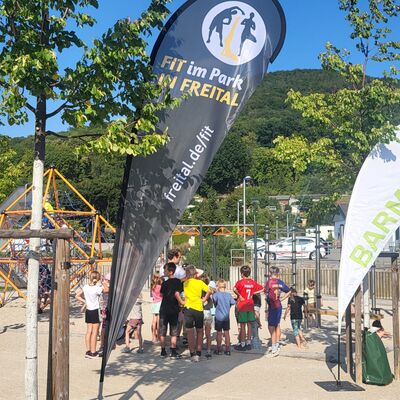 Es ist Sommer. Auf einem Spielplatz mit Kletterspinne und Hangelelementen stehen circa 15 Kinder und ein &Uuml;bungsleiter in Sportsachen. Zwischen neu gepflanzten B&auml;umen stehen Beachflags von Fit im Park und der Barmer-Krankenkasse. Im Hintergrund sind weitere Familien auf dem Spielplatz. Dahinter erhebt sich der bewaldete Windberg unter blauem Himmel.