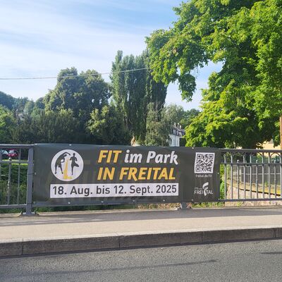 An einem Br&uuml;ckengel&auml;nder aus Metall h&auml;ngt ein Banner mit der Aufschrift Fit im Park in Freital, 18. August bis 12. September 2025. Davor ist eine Stra&szlig;e. Im Hintergrund sind B&auml;ume und H&auml;user unter blauem Himmel.
