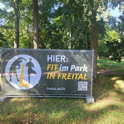 An einem Bauzaun, der in einem Park auf einer Wiese steht, h&auml;ngt ein gro&szlig;es Banner. Darauf steht Fit im Park in Freital. Im Hintergrund stehen viele alte, gro&szlig;e B&auml;ume an Wegen, die sich durch den Park ziehen.