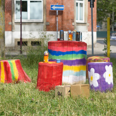 Auf einer Wiese stehen f&uuml;nf bunt bemalte Baumst&auml;mme. Sie sind verschieden gestreift, gepunktet und mit Blumen verziert. Darauf stehen Spr&uuml;hdosen. Im Hintergrund stehen H&auml;user, B&auml;ume und ein Einbahnstra&szlig;enschild an einer Stra&szlig;e.