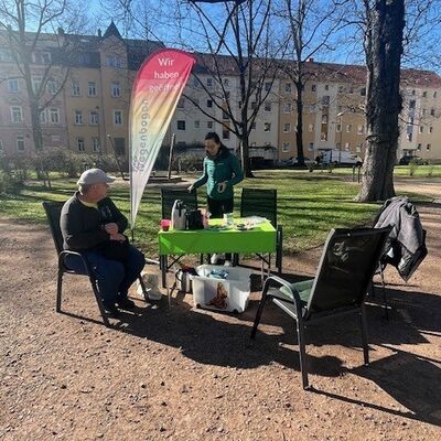 In einem Park steht im Vordergrund ein Tisch mit einer gr&uuml;nen Tischdecke. Auf dem Tisch stehen eine Teekanne und Caf&eacute;kanne. Um den Tisch herum stehen St&uuml;hle. Auf dem linken Stuhl sitzt eine Person. Eine weitere Person steht an einem Stuhl. Daneben weht eine Beachflag. Im Hintergrund ist eine Hausfassade zu erkennen.