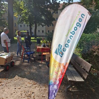 In einer Parkanlage steht im Vordergrund eine Beachflag mit dem Logo des Familienzentrums "Regenbogen" e. V.. Im Hintergrund stehen drei Personen neben einem Tisch mit St&uuml;hlen.