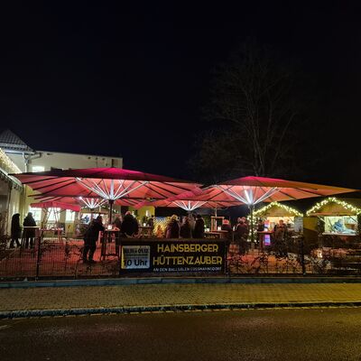 Entlang einer Stra&szlig;e stehen auf einem kleinen Platz vier rote Sonnenschirme und weihnachtlich dekorierte H&uuml;tten. Unter den Schirmen stehen Personen an Stehtischen. Links wird der Platz durch eine Hauswand begrenzt. Es ist dunkel. Die Weihnachtslichter leuchten.