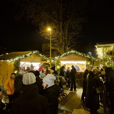 Auf einem kleinen Platz stehen im Hintergrund zwei weihnachtlich geschm&uuml;ckte H&uuml;tten. Es ist dunkel. Die Lichter leuchten weihnachtlich. Im Vordergrund stehen viele Personen.