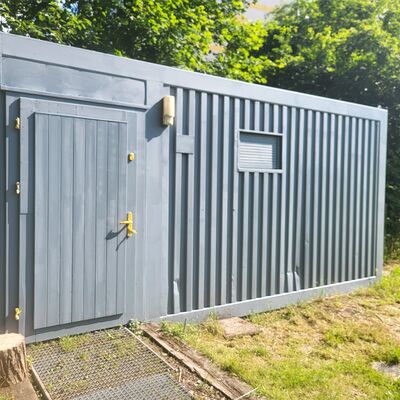 Auf einer Wiese steht ein hellblauer Container. Der Container hat auf der linken Seite eine T&uuml;r. Auf der rechten Seite befindet sich ein schmales Fenster. Im Hintergrund sind B&auml;ume.