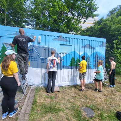 Auf einer Wiese steht ein blauer Container. Der Container wird von f&uuml;nf Kindern bunt angespr&uuml;ht. Ein Mann steht auf einer Leiter. Als Bild entsteht ein graues Haus mit B&auml;umen, Wolken und Bergen im Hintergrund.