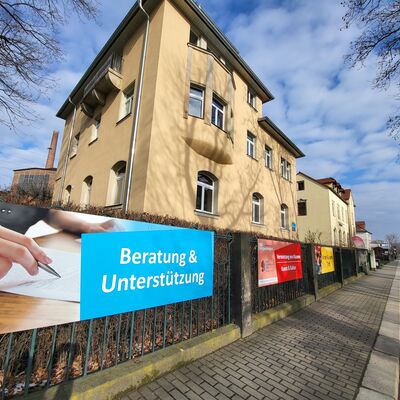 An einem Zaun an einem Gehweg h&auml;ngen Werbeplakate. Auf dem vordersten Werbeplakat steht "Beratung und Unterst&uuml;tzung". Hinter dem Zaun ist ein gro&szlig;es Wohnhaus.