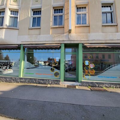 Am unteren Teil einer Geb&auml;udefassade befindet sich ein gr&uuml;nes Schaufenster. Darauf steht "Kreativ K&ouml;pfe Freital, Tanz, Sport und mehr". Im Schaufenster befinden sich mehrere bunte Kreise, in denen Aush&auml;nge ausgestellt werden.
