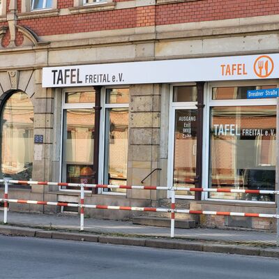 Das Schaufenster der Tafel Freital e. V. befindet sich im unteren Teil eines Mehrfamilienhauses. Davor ist ein gepflasterter Gehweg hinter einem gestreiften Zaun.