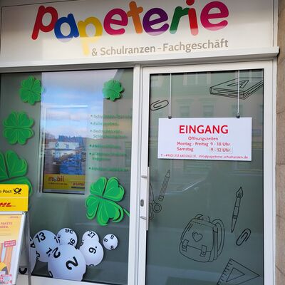 An einem Schaufenster mit einer T&uuml;r h&auml;ngt ein Schild mit den &Ouml;ffnungszeiten des Gesch&auml;ftes. Auf dem Schaufenster sind Kleebl&auml;tter und Lottokugeln. Dar&uuml;ber steht Papeterie und Schulranzen Fachgesch&auml;ft. Vor dem Gesch&auml;ft steht ein Aufsteller.