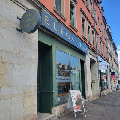 In einer Geb&auml;udefassade befindet sich das Schaufenster eines Friseurs. Im Fenster sind eine Schere und die Aufschrift Friseur, Kosmetik, Fu&szlig;pflege zusehen. Vor dem Gesch&auml;ft steht ein Aufsteller mit Werbung. Im Hintergrund sind entlang der Stra&szlig;e weitere Schaufenster und Aufsteller zu erkennen.