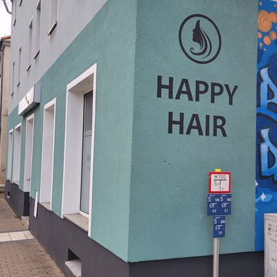 An einer t&uuml;rkis bemalten Hausecke befindet sich der Friseur Happy Hair. Auf der t&uuml;rkisen Fl&auml;che ist das Logo des Friseurs, ein Kopf mit langen Haaren in einem Kreis, zu sehen.