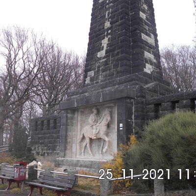 Im Mittelpunkt steht ein aus Sandstein gebautes Denkmal in Form eines Obelisken mit Reiterrelief. Hinter dem Denkmal sind kahle B&auml;ume. Vor dem Denkmal stehen zwei B&auml;nke und ein M&uuml;lleimer.
